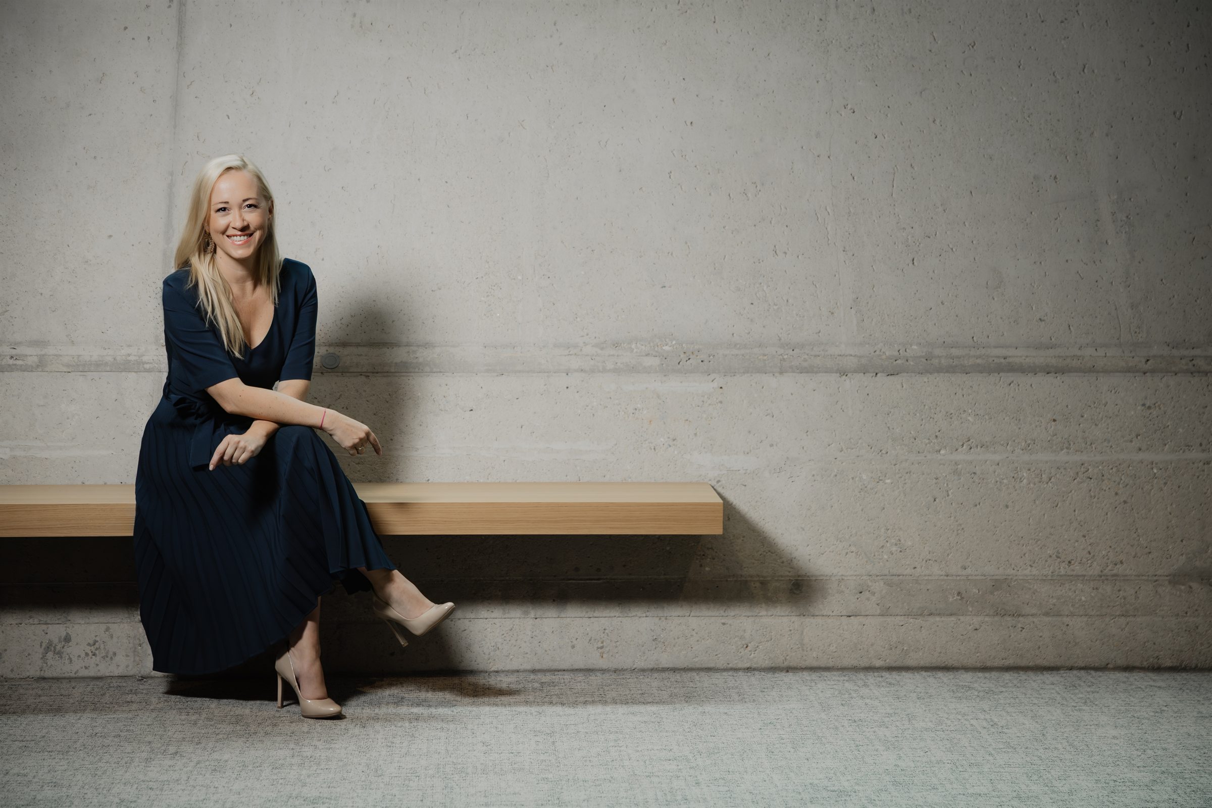 Sabine Kronberger sitzt lächelnd auf einer Holzbank vor einer Betonwand